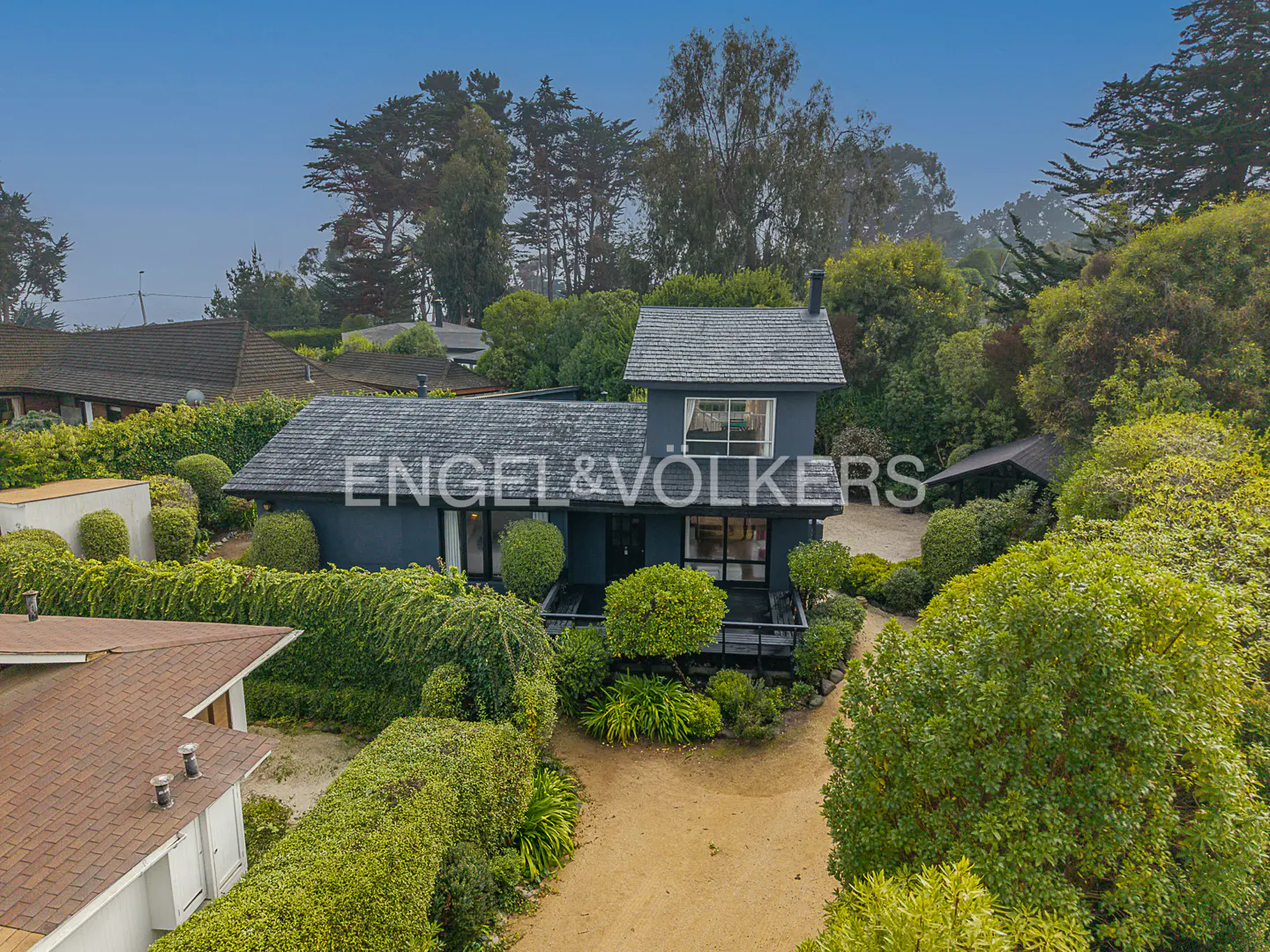 Aerial view of a dark blue two-story house with a gray roof, surrounded by green trees and bushes. "Engel & Völkers" is overlaid on the image.