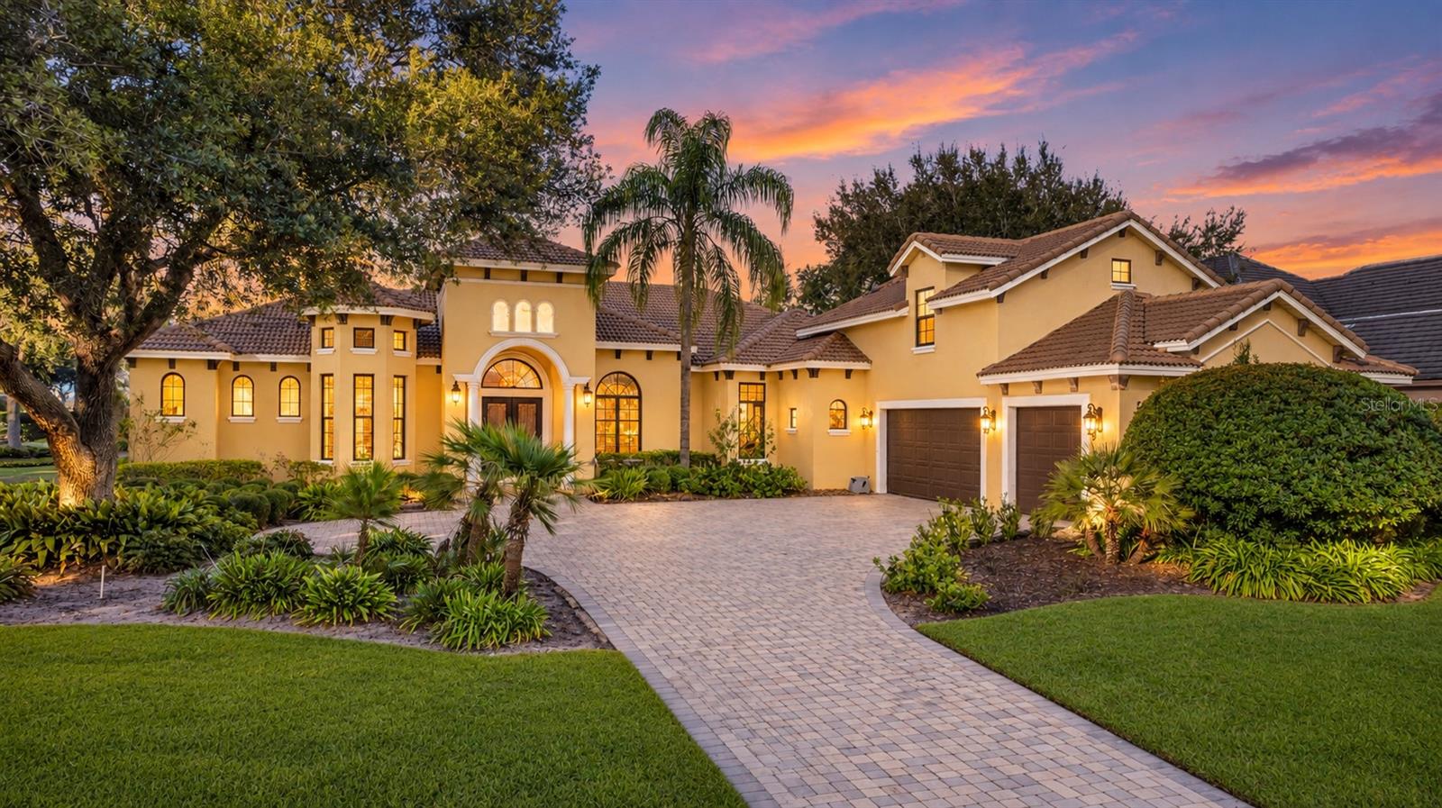 Yellow Mediterranean-style home with a paver driveway and lush landscaping at sunset. [AI-generated text]