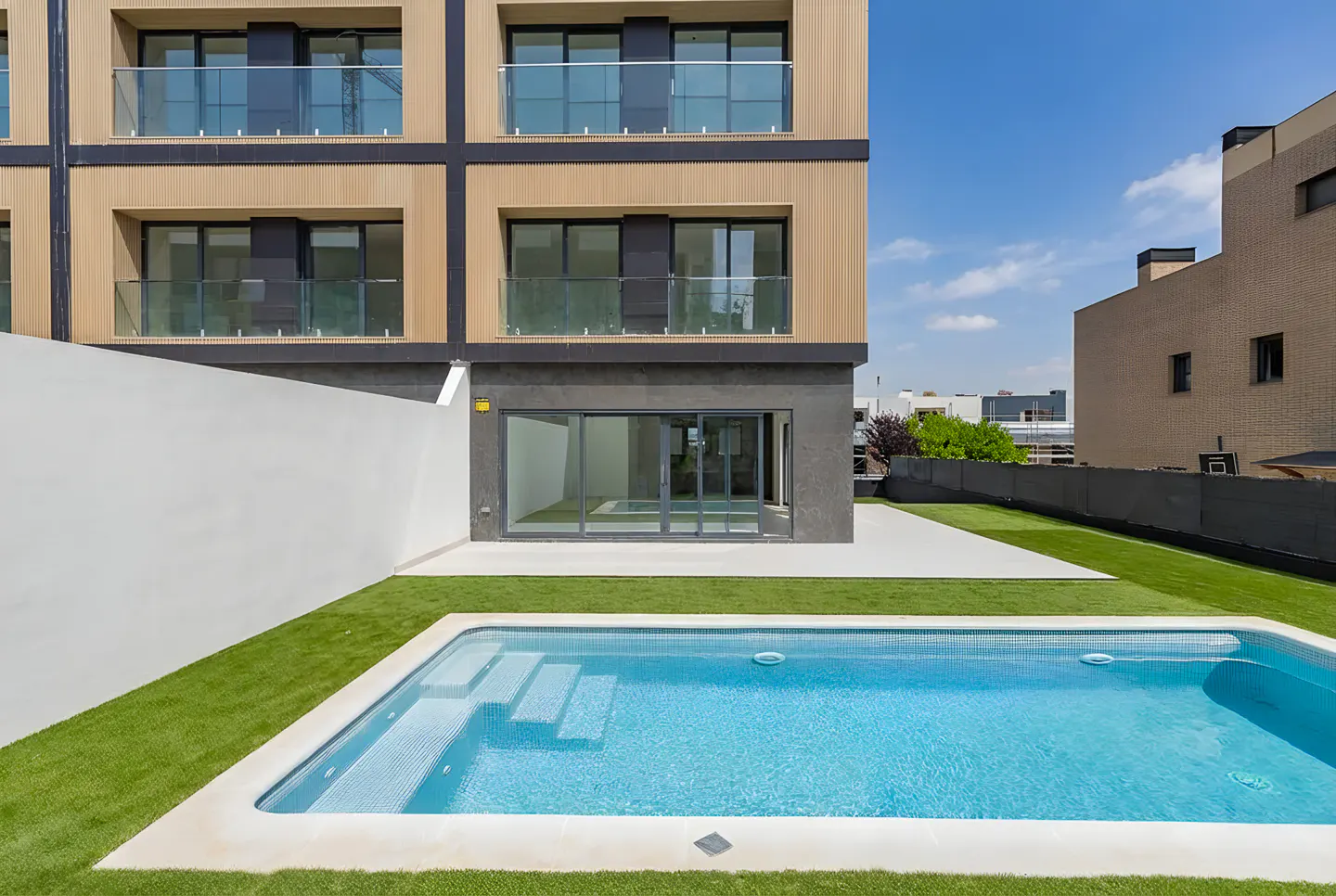 Modern building with a blue swimming pool and green lawn. The building has balconies and large sliding glass doors.