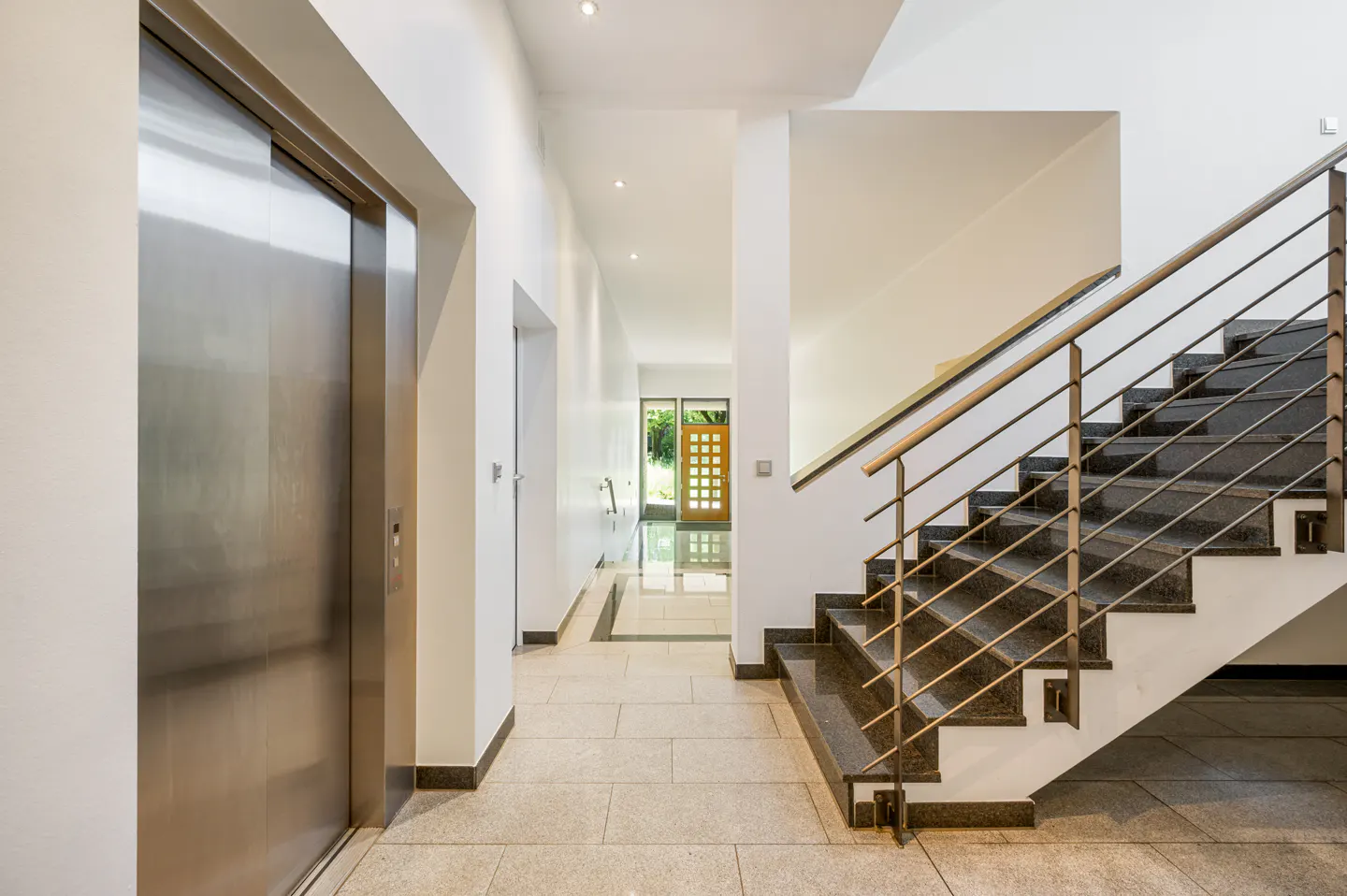 Modern home interior with a stainless steel elevator, marble stairs with metal railings, and a hallway leading to a wooden door.