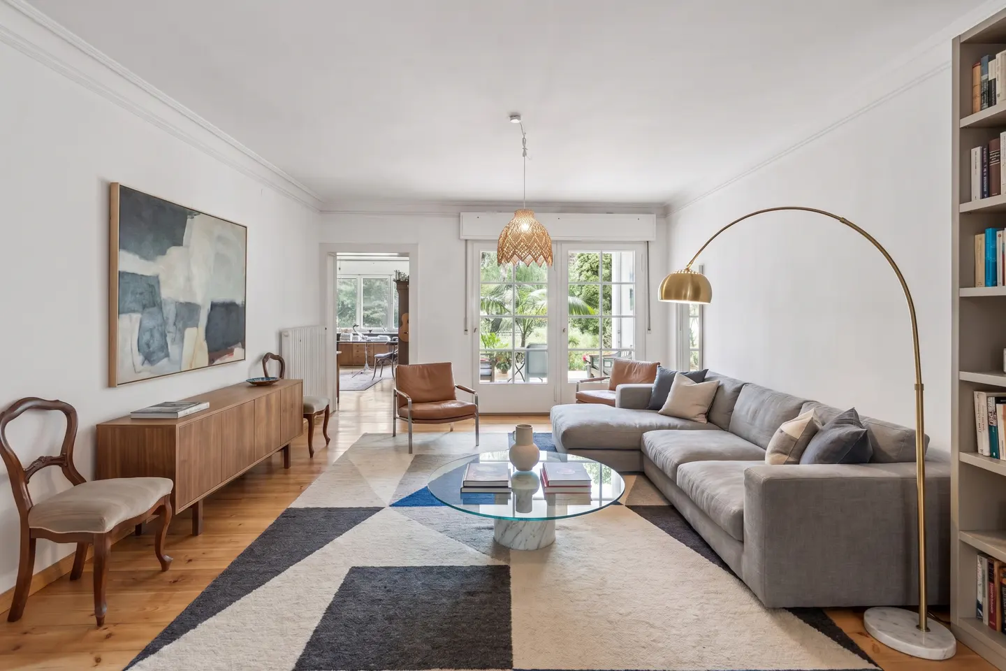 Bright living room with a gray sectional sofa, a glass coffee table, and a geometric rug. A gold arc lamp and a bookcase are on the right.