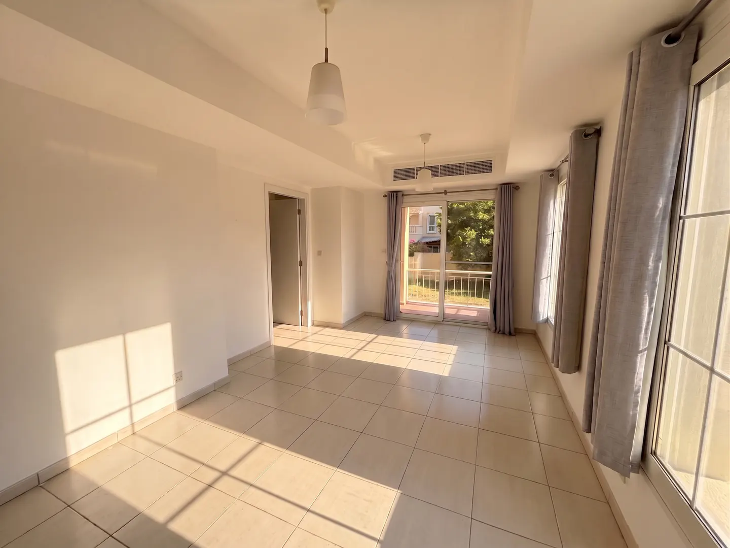 Bright, empty room with beige tile floor, white walls, and gray curtains. A glass door leads to a balcony with a view of trees.