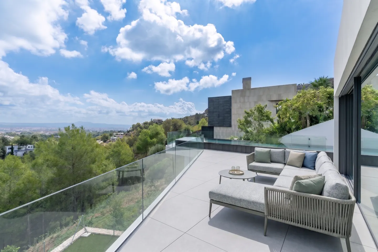 Outdoor patio with gray sectional sofa, glass railing, and city view under a blue sky with white clouds.