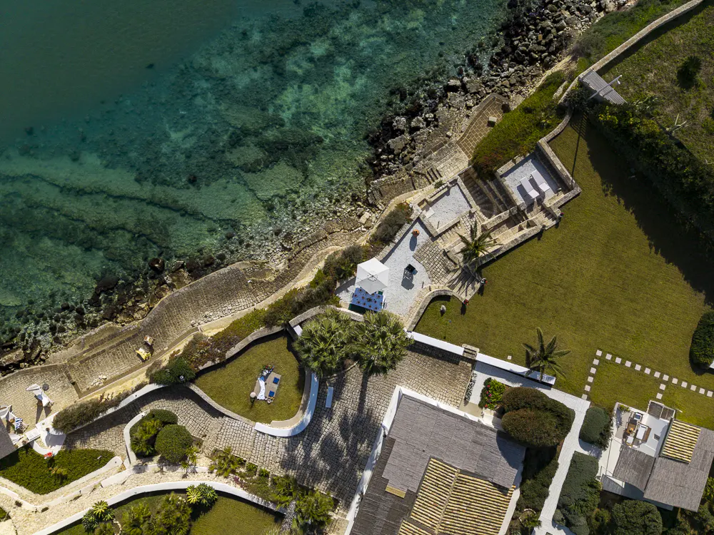 Aerial view of a coastal property with turquoise water, stone terraces, green lawns, and buildings with light-colored roofs.