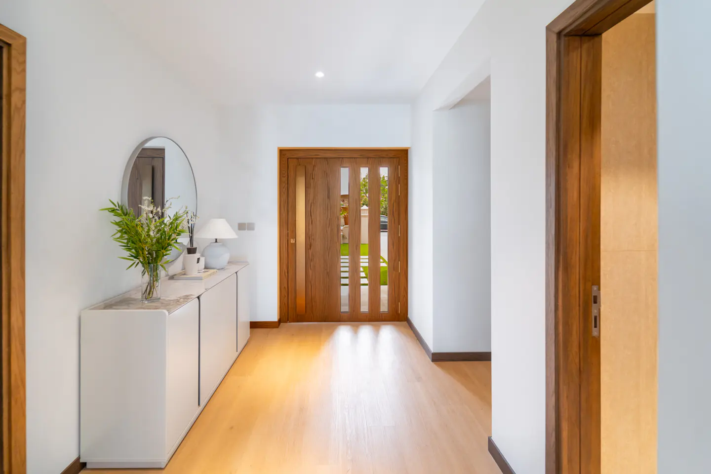 Bright foyer with wood floors, white walls, and a wood front door with vertical glass panels. A white cabinet with decor and a round mirror sits to the left.