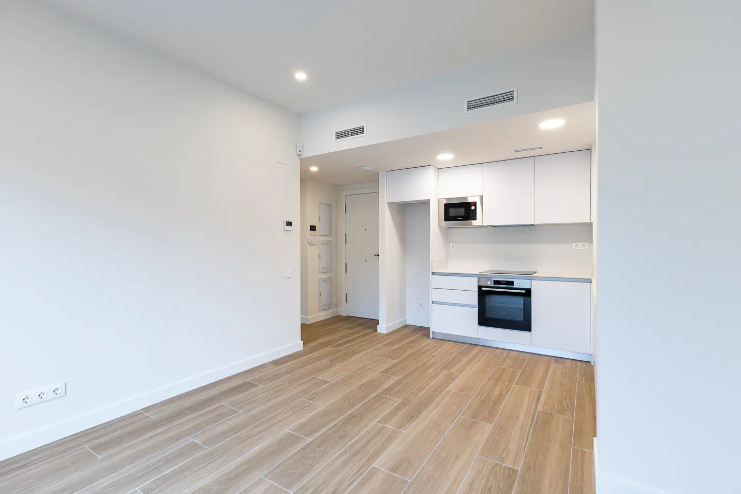 Bright, modern apartment interior with white walls, wood-look tile flooring, and a compact kitchen featuring white cabinets and appliances.