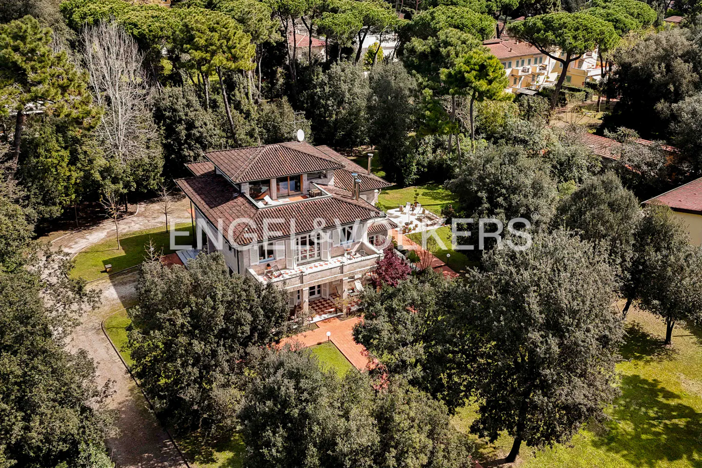 Aerial view of a two-story house with a red tile roof, surrounded by green trees and lawns. The house has a balcony and a patio.