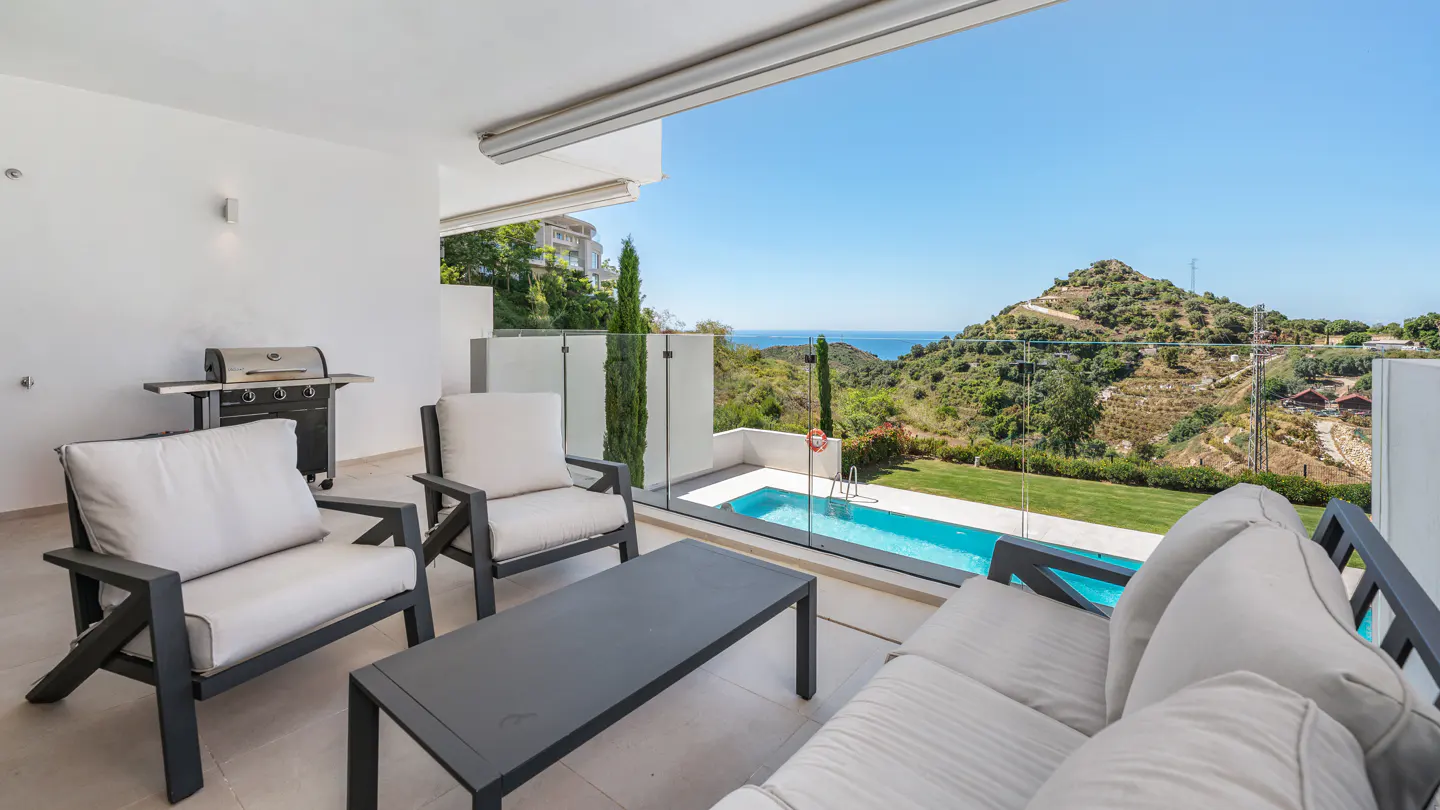 Outdoor patio with seating, grill, and pool overlooking a green hillside and blue ocean under a clear sky.