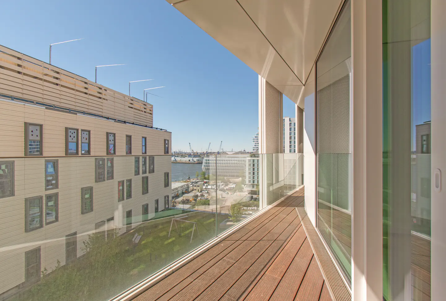 Balcony view with wood floor, glass railing, and city view. Buildings, water, and blue sky visible.