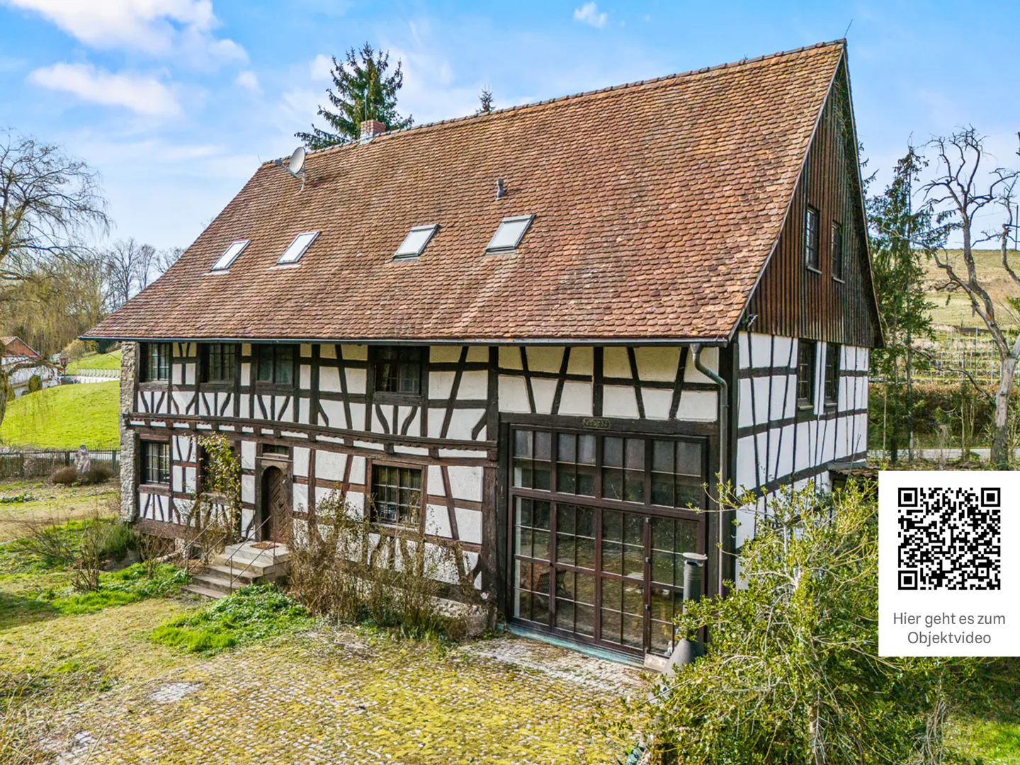 Traditional half-timbered house with a brown tiled roof, large windows, and a cobblestone yard under a blue sky.