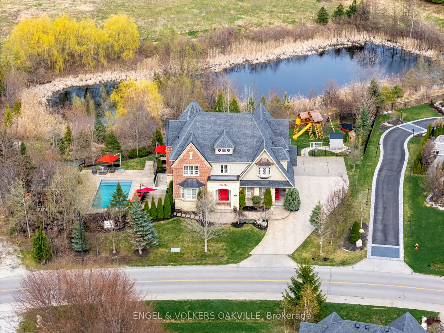 Aerial view of a large, multi-story house with a pool, pond, and playground in the backyard. The house has a red front door.