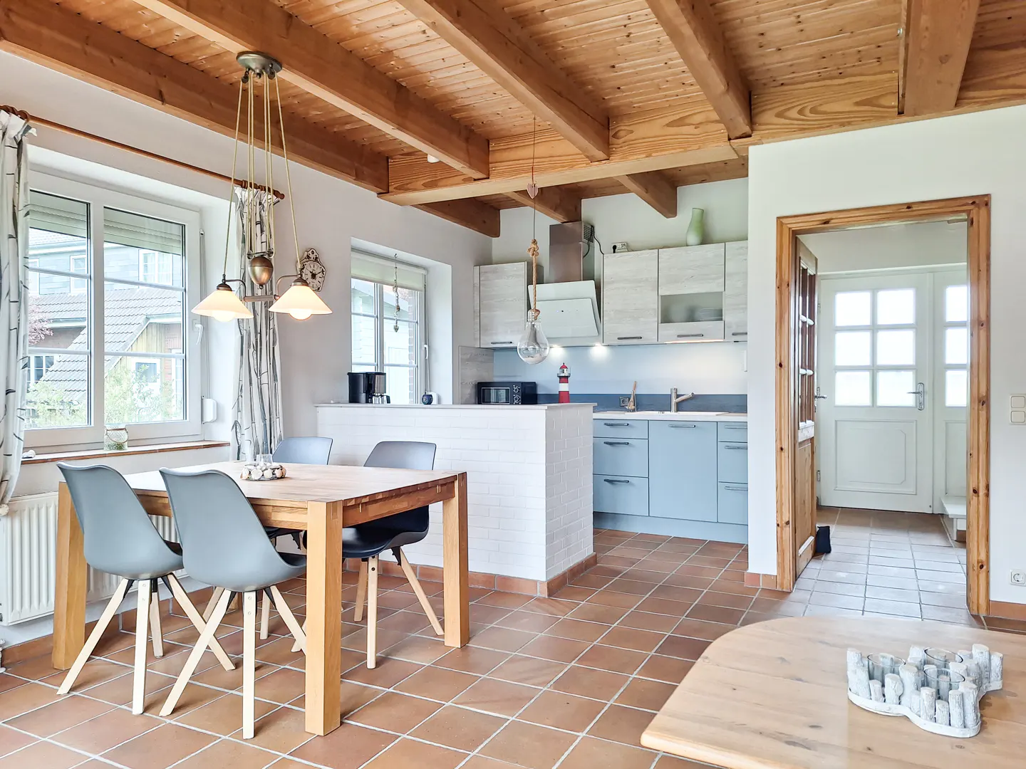 A bright kitchen and dining area with a wooden table, gray chairs, and blue cabinets. The ceiling has exposed wooden beams.