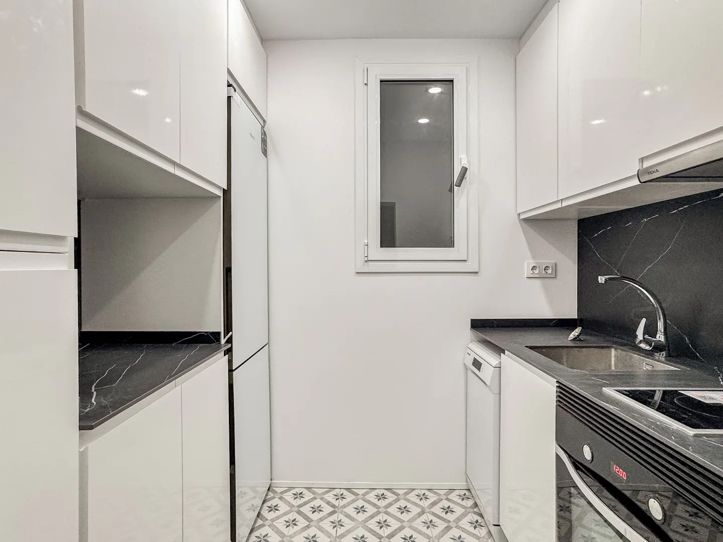 A modern, narrow kitchen with white cabinets, black marble countertops, and patterned tile flooring. A window provides natural light.