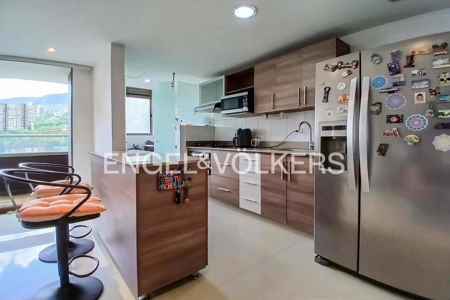 Modern kitchen with wood cabinets, stainless steel refrigerator, island with two bar stools, and a view of city buildings.