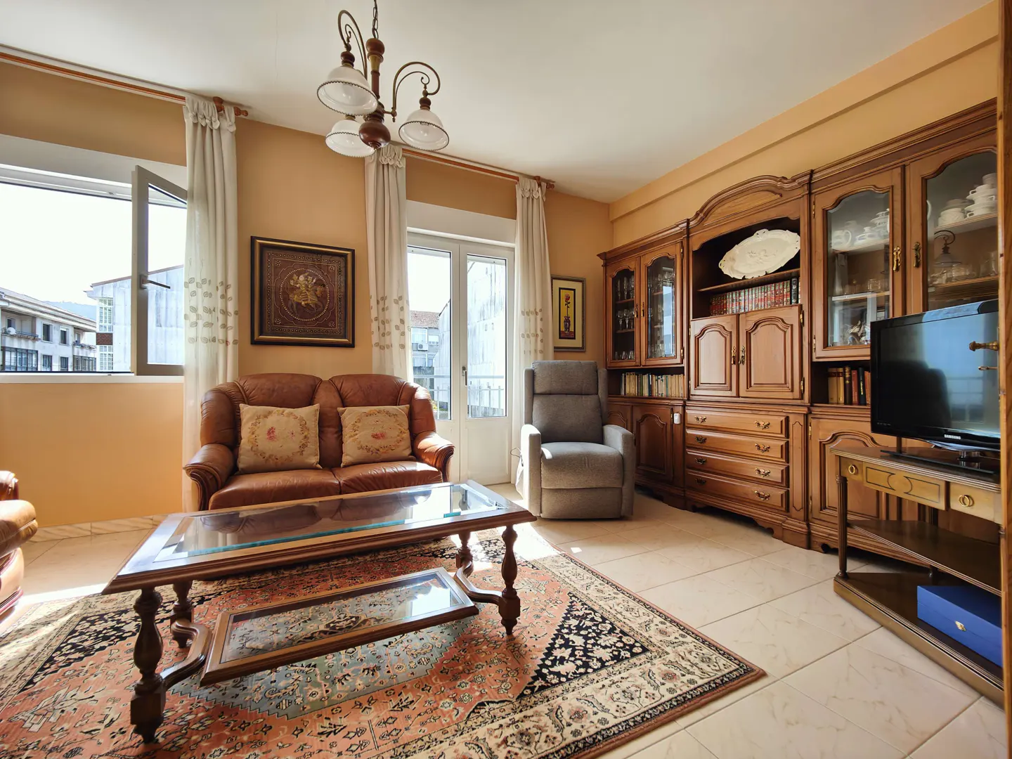 Living room with brown leather sofa, glass coffee table, patterned rug, wooden cabinet, and gray recliner. Light walls and tile floor.