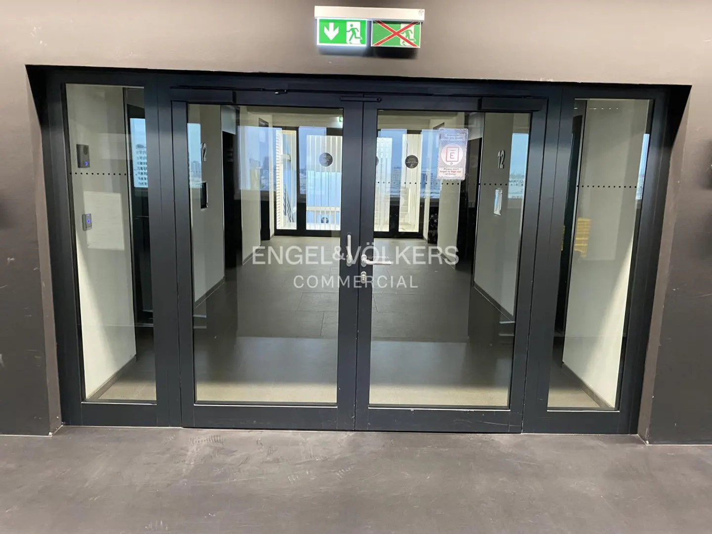 Glass doors with black frames lead to an Engel & Volkers Commercial office. An exit sign is above the doors.