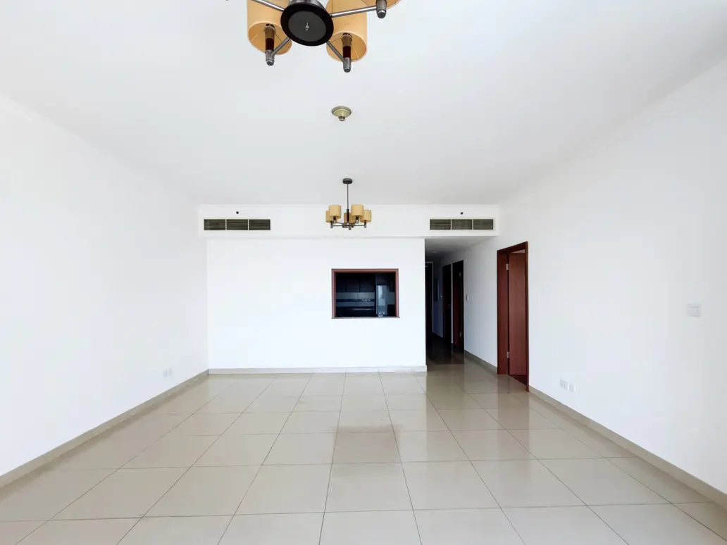 Empty living room with white walls, beige tile floor, and two chandeliers. A doorway leads to a hallway with dark doors.