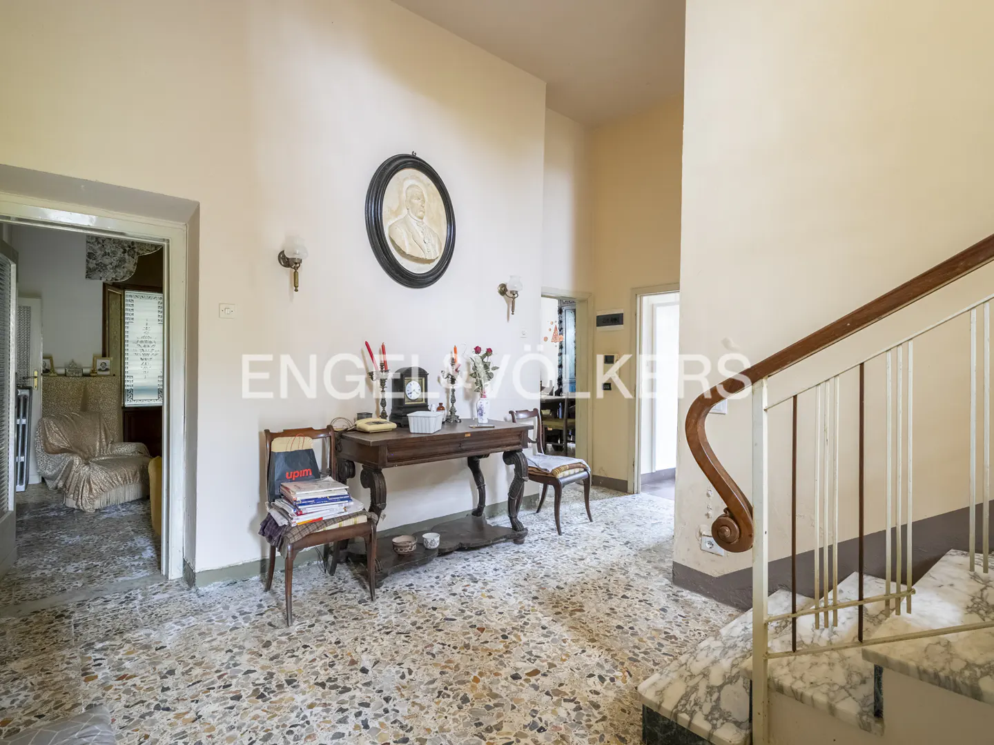 Foyer with terrazzo floor, antique table, portrait, and staircase with marble steps and white railing.