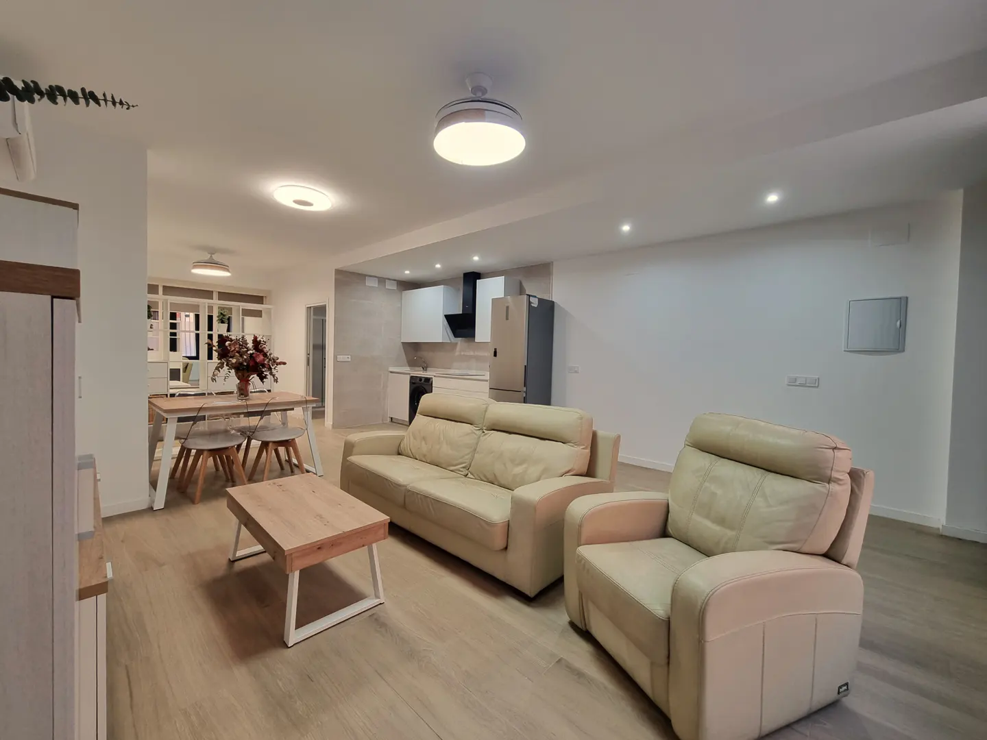 Open-concept living space with a beige sofa, armchair, and wood coffee table. Dining area and kitchen visible in the background.