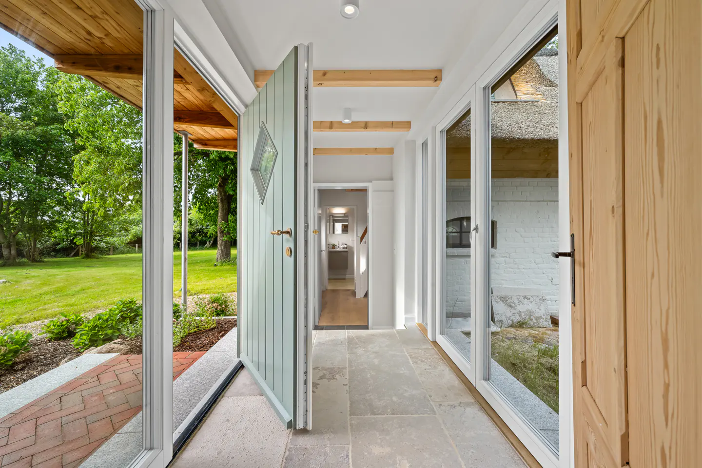 A bright hallway with stone floors, a sage green door, and glass walls looking out onto a lush green garden.