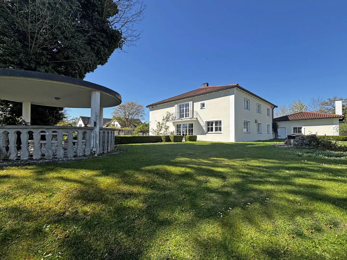 Exterior view of a two-story white house with a red tile roof, a gazebo, and a green lawn on a sunny day.