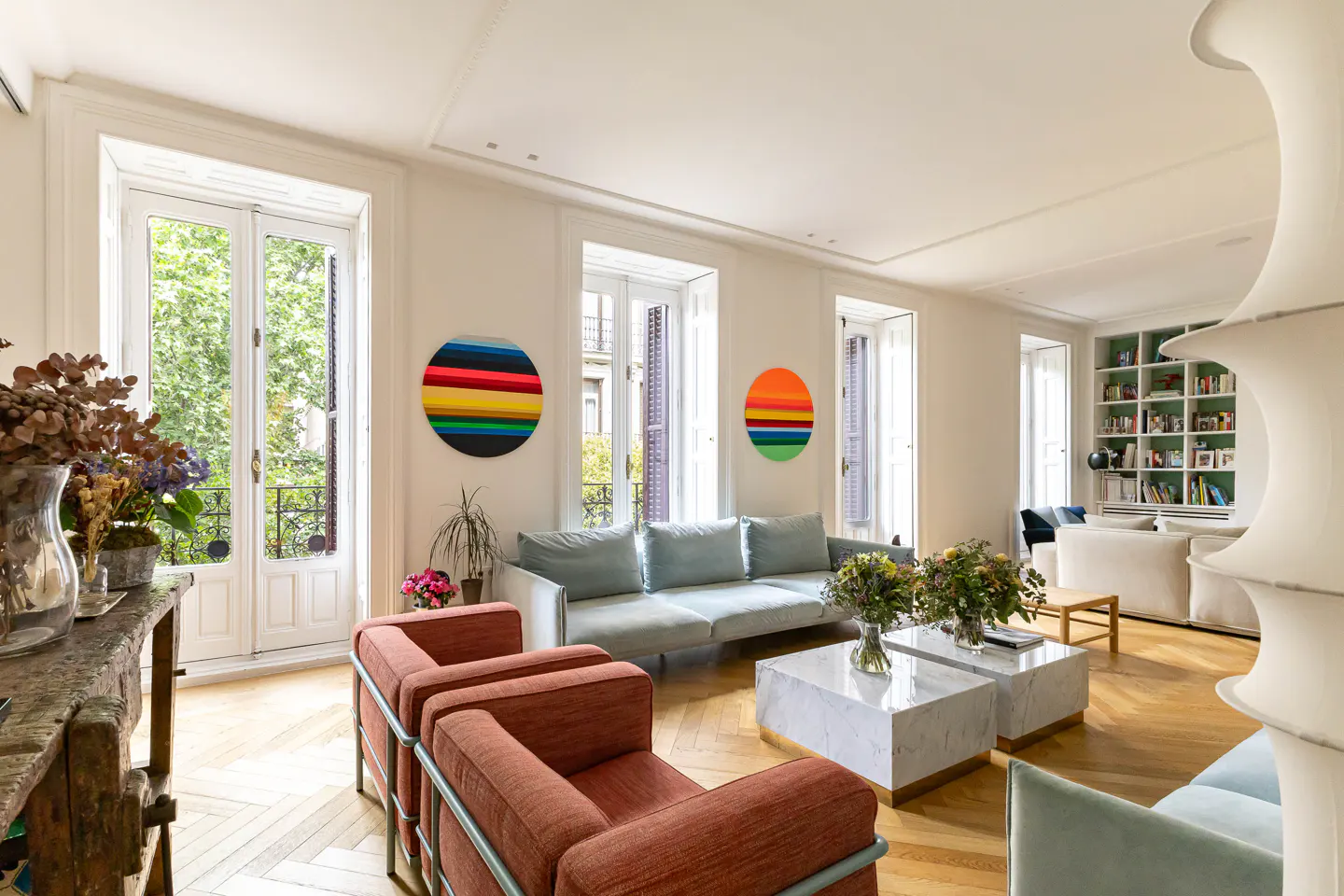Bright living room with parquet floors, white walls, and colorful art. Two orange chairs face a light blue sofa and marble coffee tables. French doors open to a balcony.
