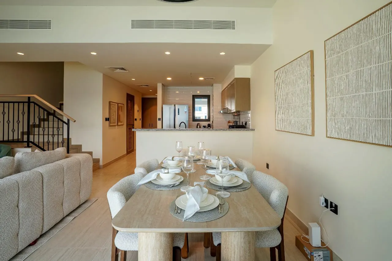 A dining area with a light wood table set for six, surrounded by white chairs. Artwork hangs on the wall, and a kitchen is visible in the background.