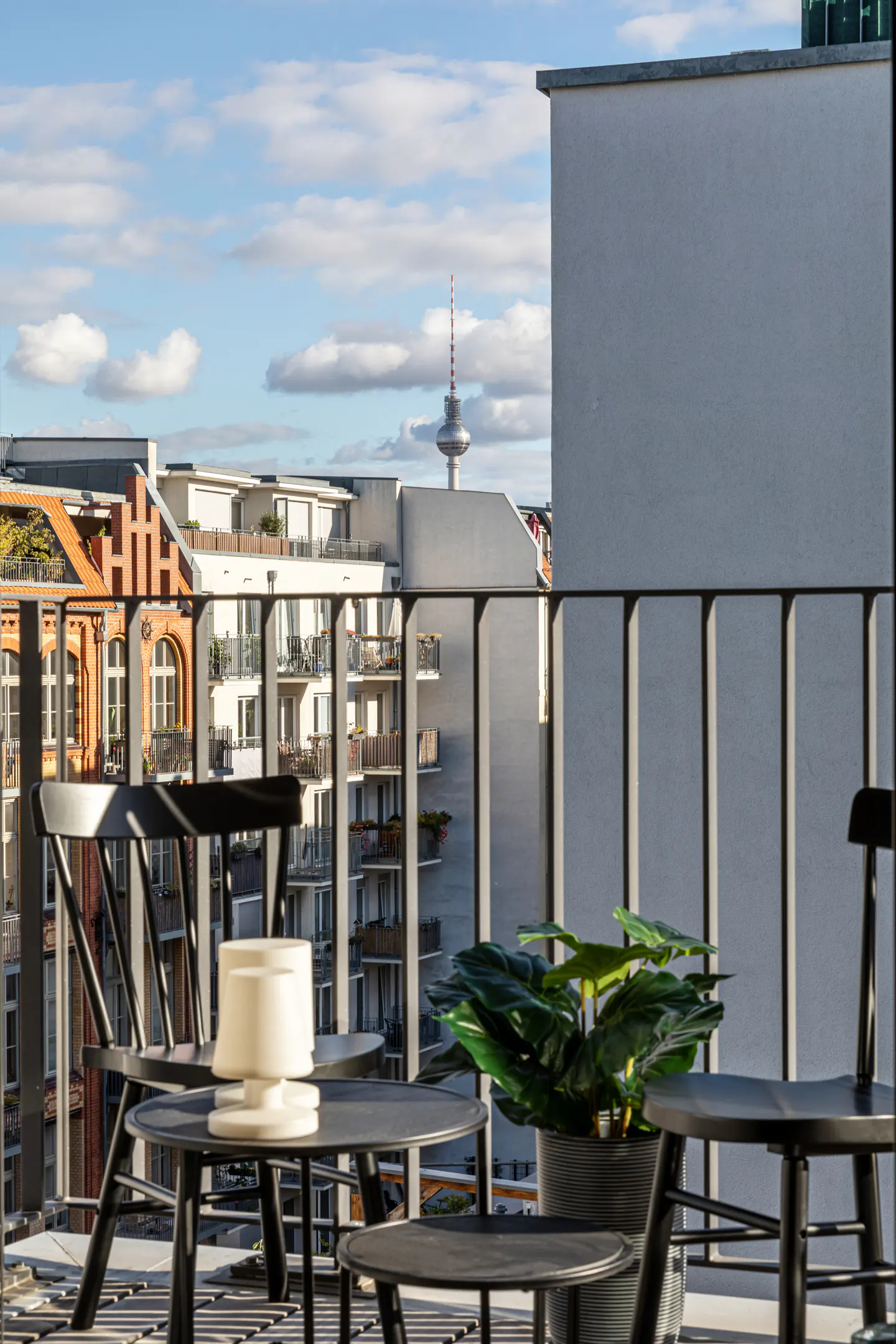 Balcony view with black chairs, table, lamp, and plant. City buildings and the Berlin TV Tower are visible in the background.