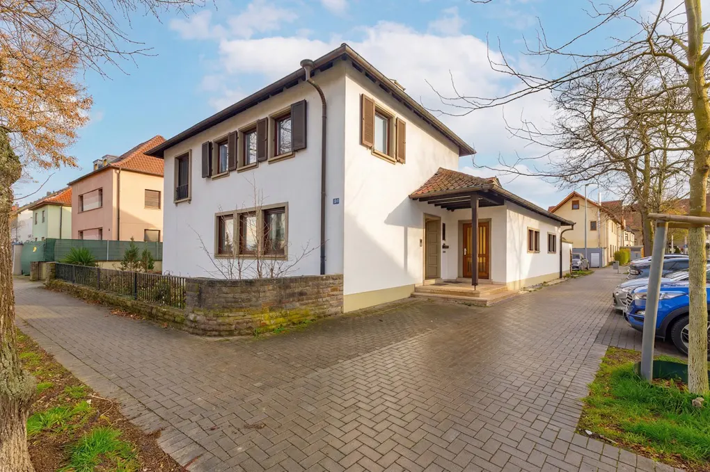 Exterior view of a two-story white house with brown shutters and a tiled roof, on a brick-paved street.