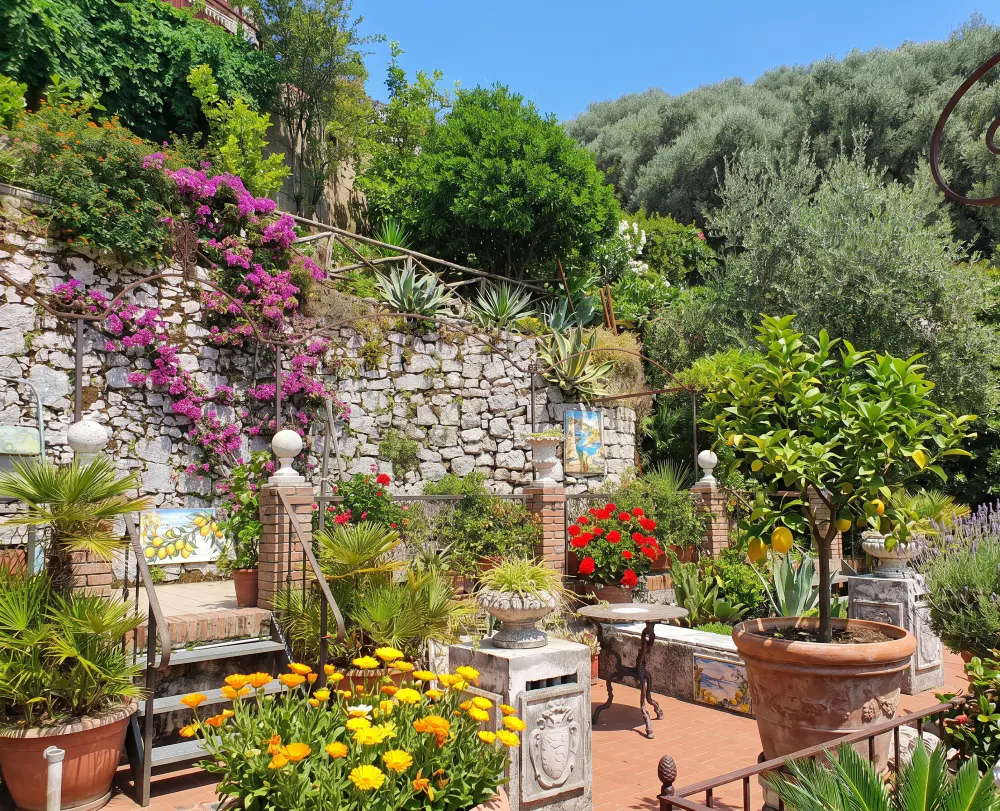 A vibrant garden scene with stone walls, colorful flowers, potted plants, and a small table on a sunny day.