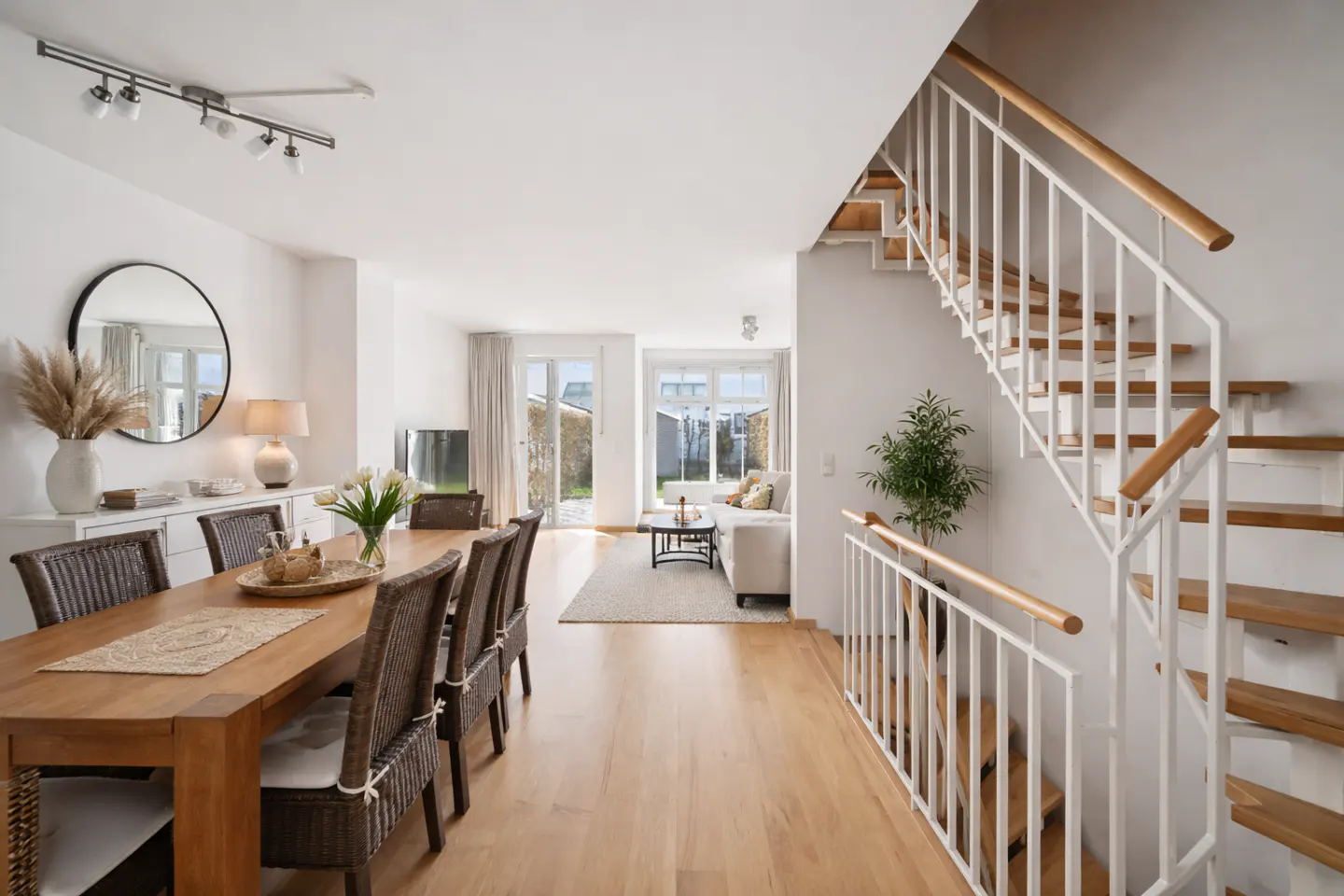 Bright, open-concept home interior with wood floors, dining table, white walls, and a staircase with white railings. A living area is visible in the background.