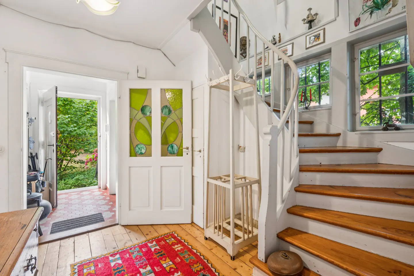 Entryway with white walls, wood floors, and a red rug. A white staircase curves up to a window. A door with green stained glass is to the left.