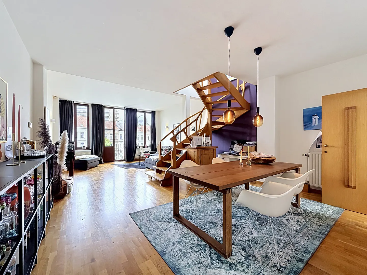 Open-plan living area with a wooden dining table, white chairs, a blue patterned rug, and a wooden staircase.