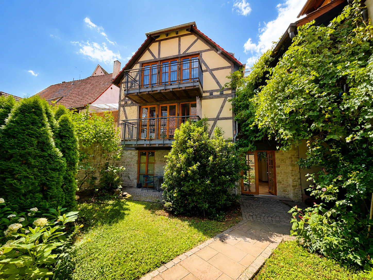 A half-timbered house with two balconies overlooks a lush green garden with a stone path under a bright blue sky. [AI-generated text]