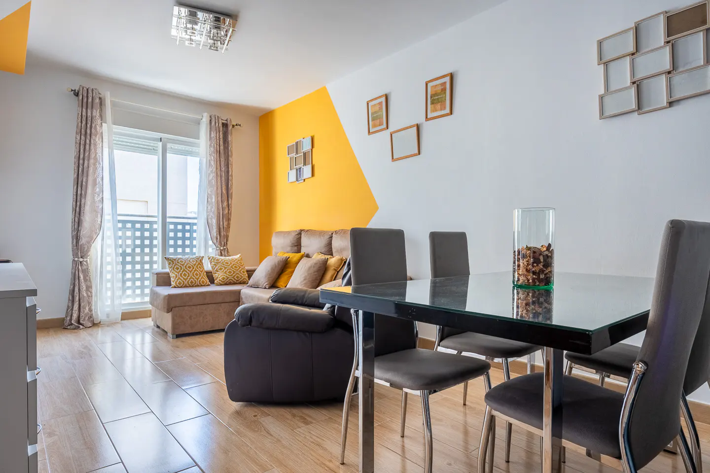 Bright living room with a brown sectional sofa, a glass-top dining table with four chairs, and a yellow accent wall.