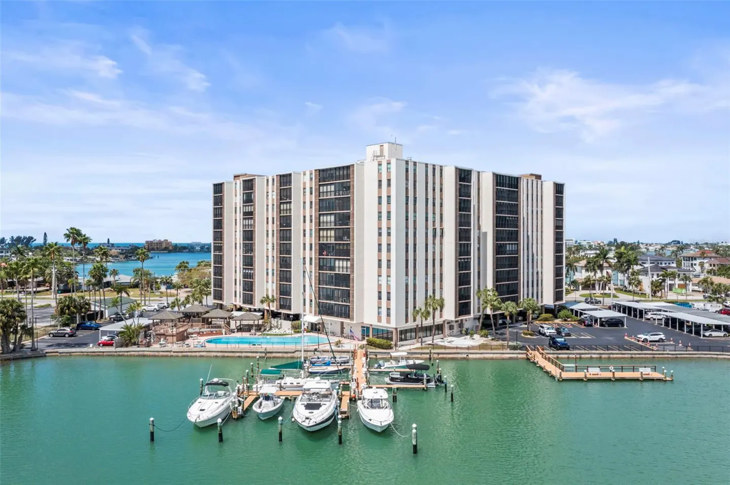 Aerial view of a white condo building with a marina, pool, and parking lot on a sunny day.