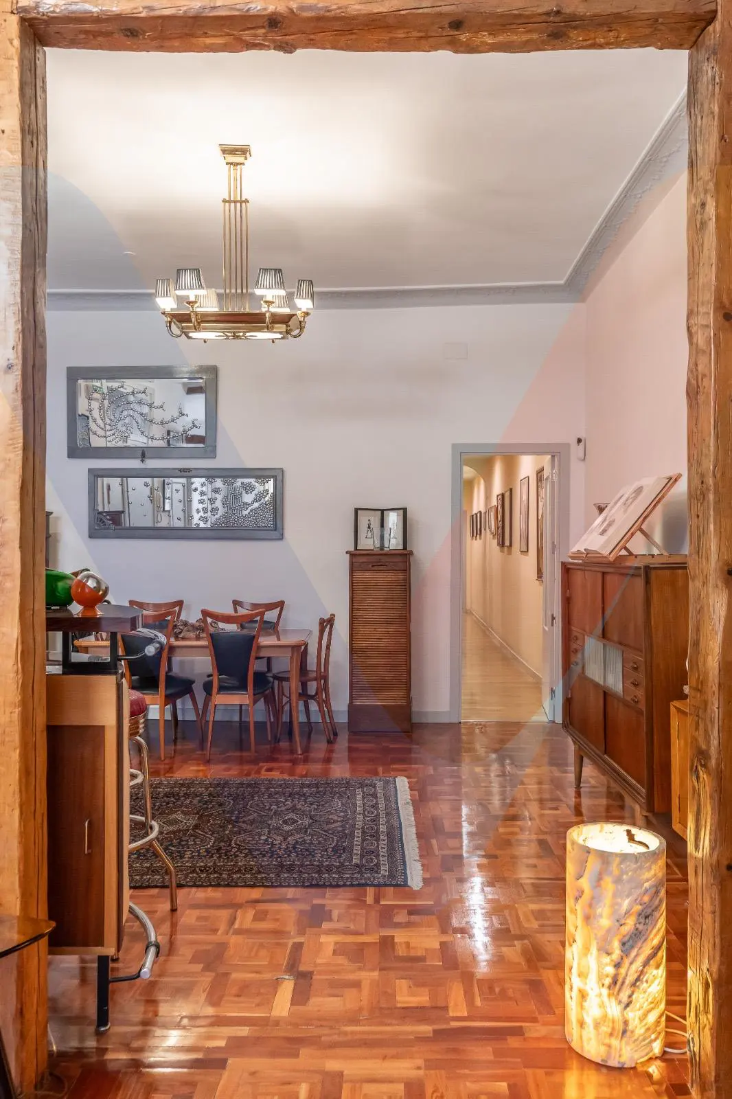 A dining room with wooden floors, a table with chairs, and a hallway with art on the walls. A chandelier hangs above the table.