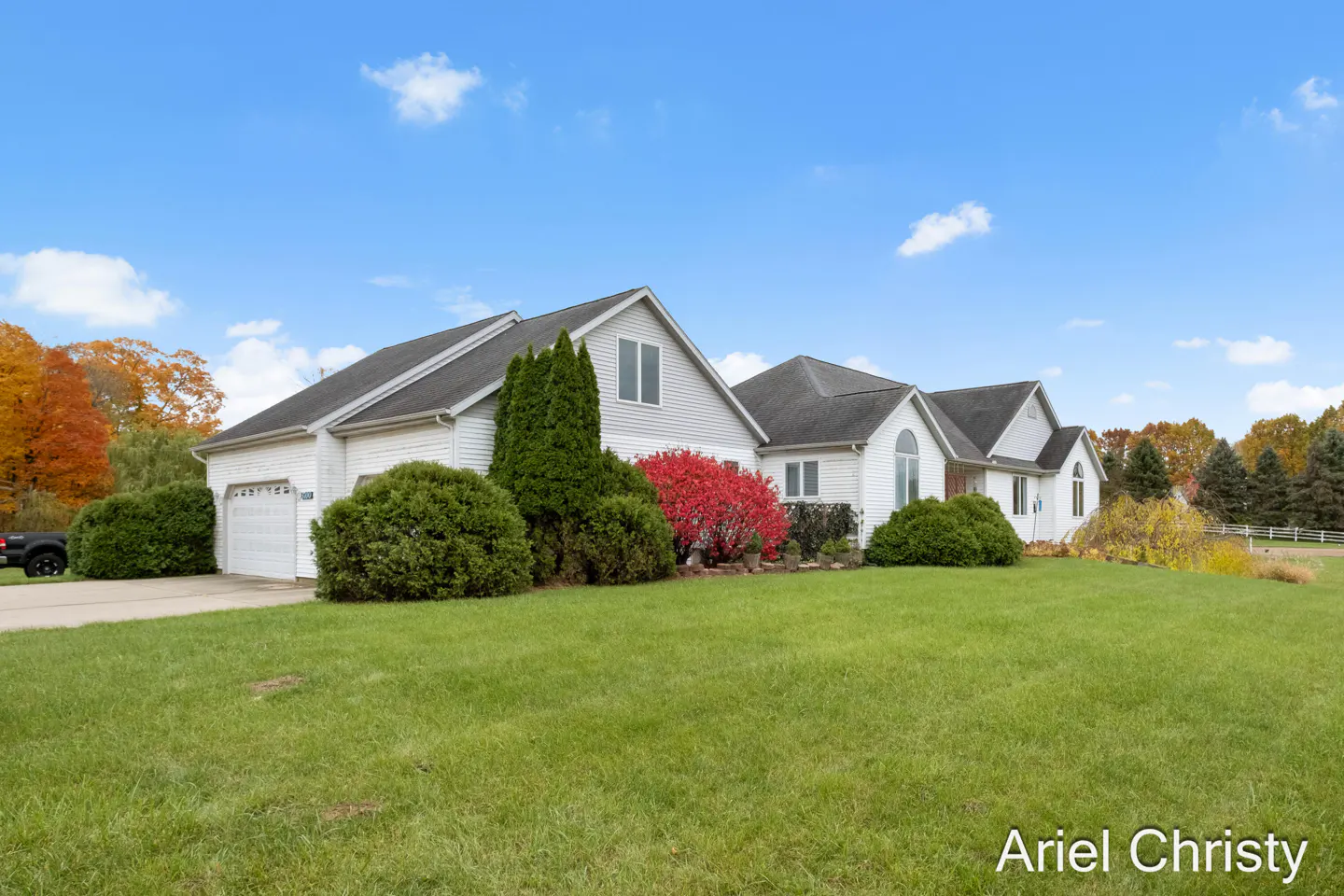 A white, multi-sectioned house with a gray roof sits on a green lawn under a blue sky. Bushes and trees surround the house.