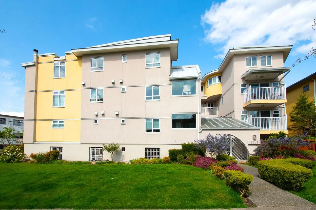 Exterior view of a modern, multi-story apartment building with beige and yellow walls, a green lawn, and a blue sky.