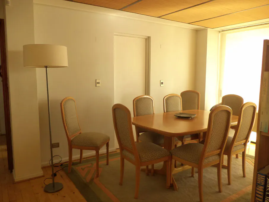 A dining room with a wooden table, eight chairs, a floor lamp, and a patterned rug. The walls are white, and the ceiling is a light brown.