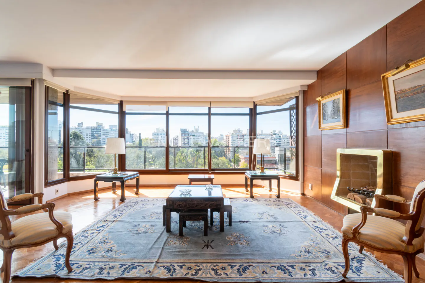 Bright living room with wood floors, a blue rug, and a wall of windows overlooking a city park. A fireplace is on the right.