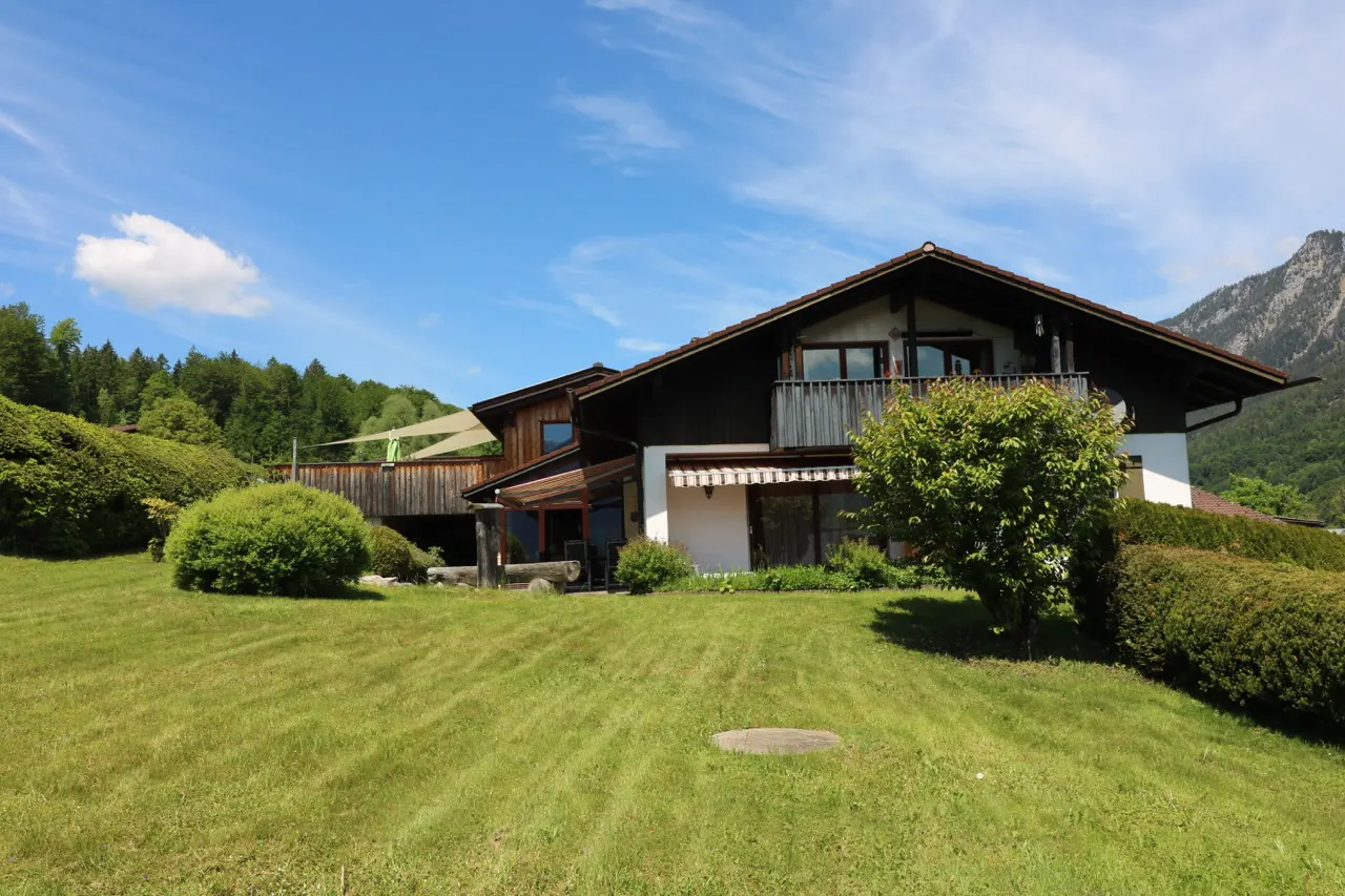 A two-story house with dark wood siding and a balcony sits on a lush green lawn against a backdrop of mountains and sky.