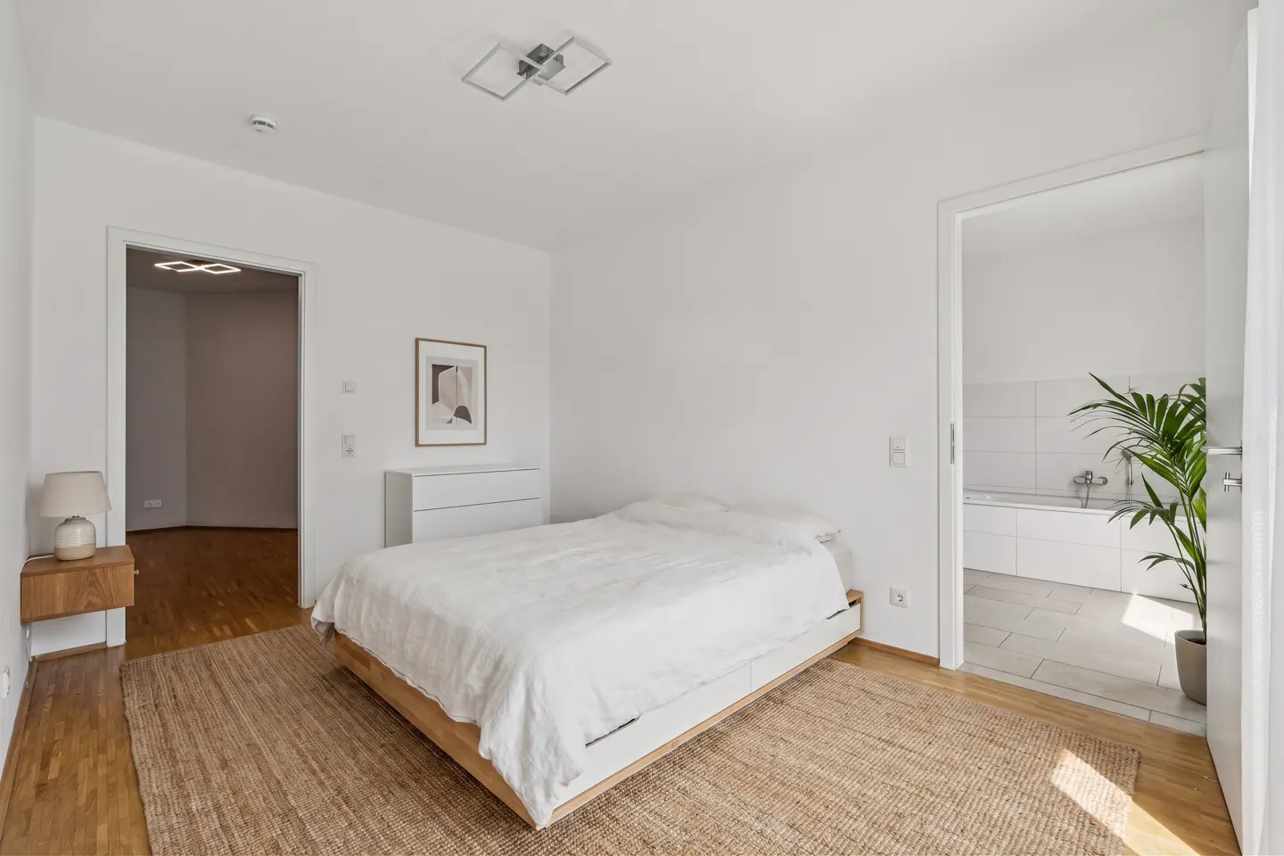 Minimalist bedroom with a white bed on a jute rug, wooden nightstand, and an open doorway leading to a tiled bathroom.