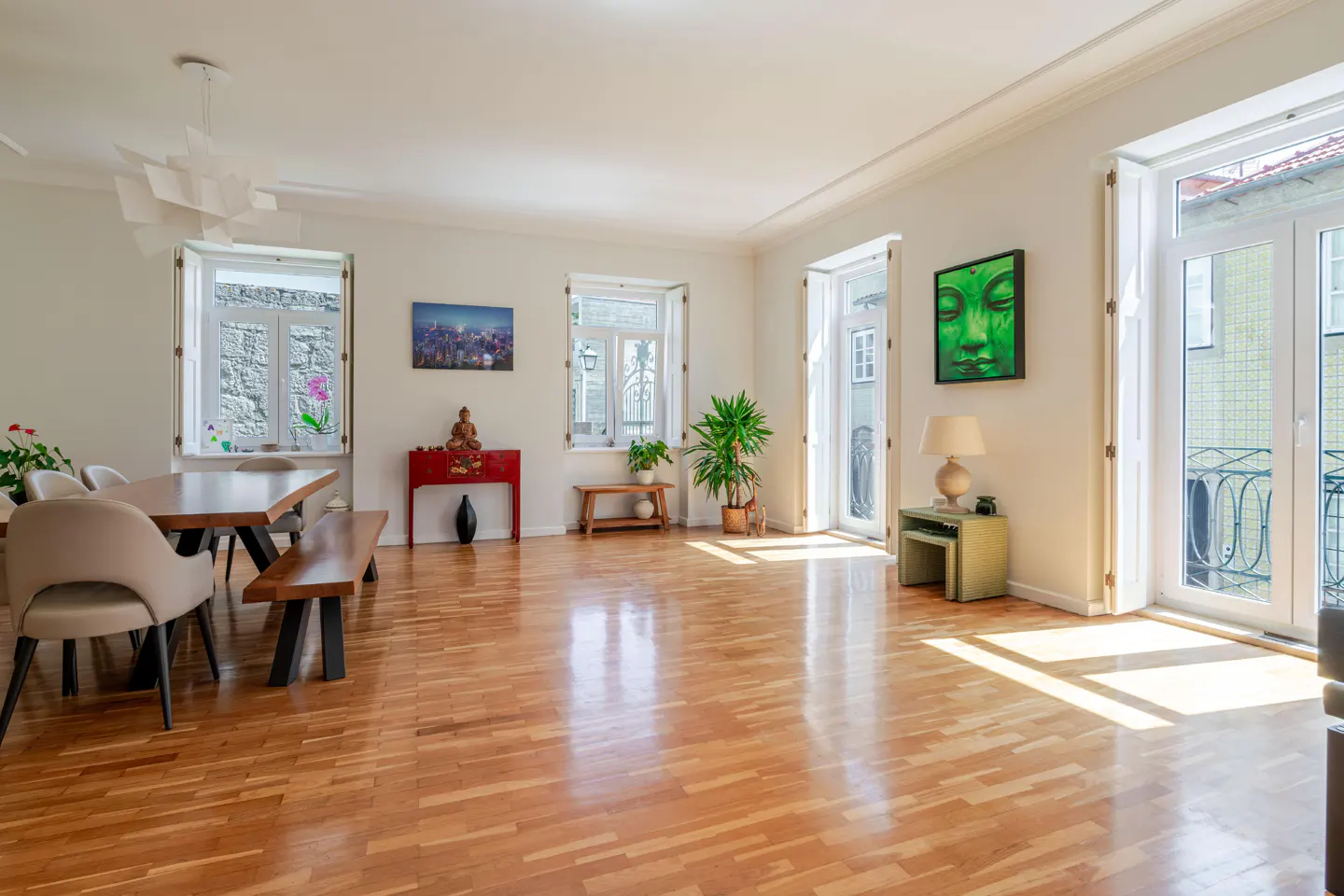 Bright, airy living room with wood floors, white walls, and large windows. Dining table, bench, and chairs are on the left. Artwork and plants add color.