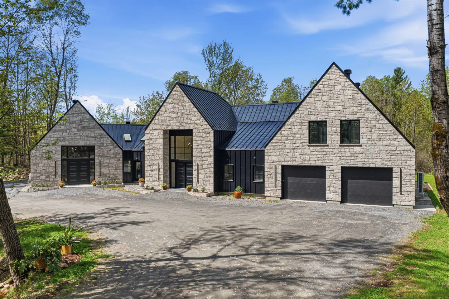 Modern stone house with black trim and a metal roof on a gravel driveway, surrounded by trees.
