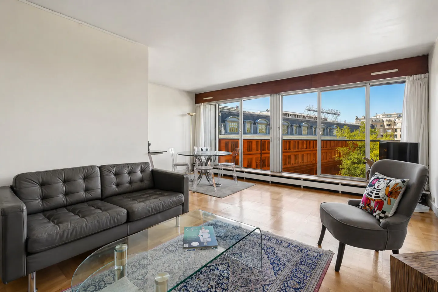 A bright living room with a black leather sofa, a glass coffee table, and a gray armchair with a colorful pillow. Large windows offer a city view.