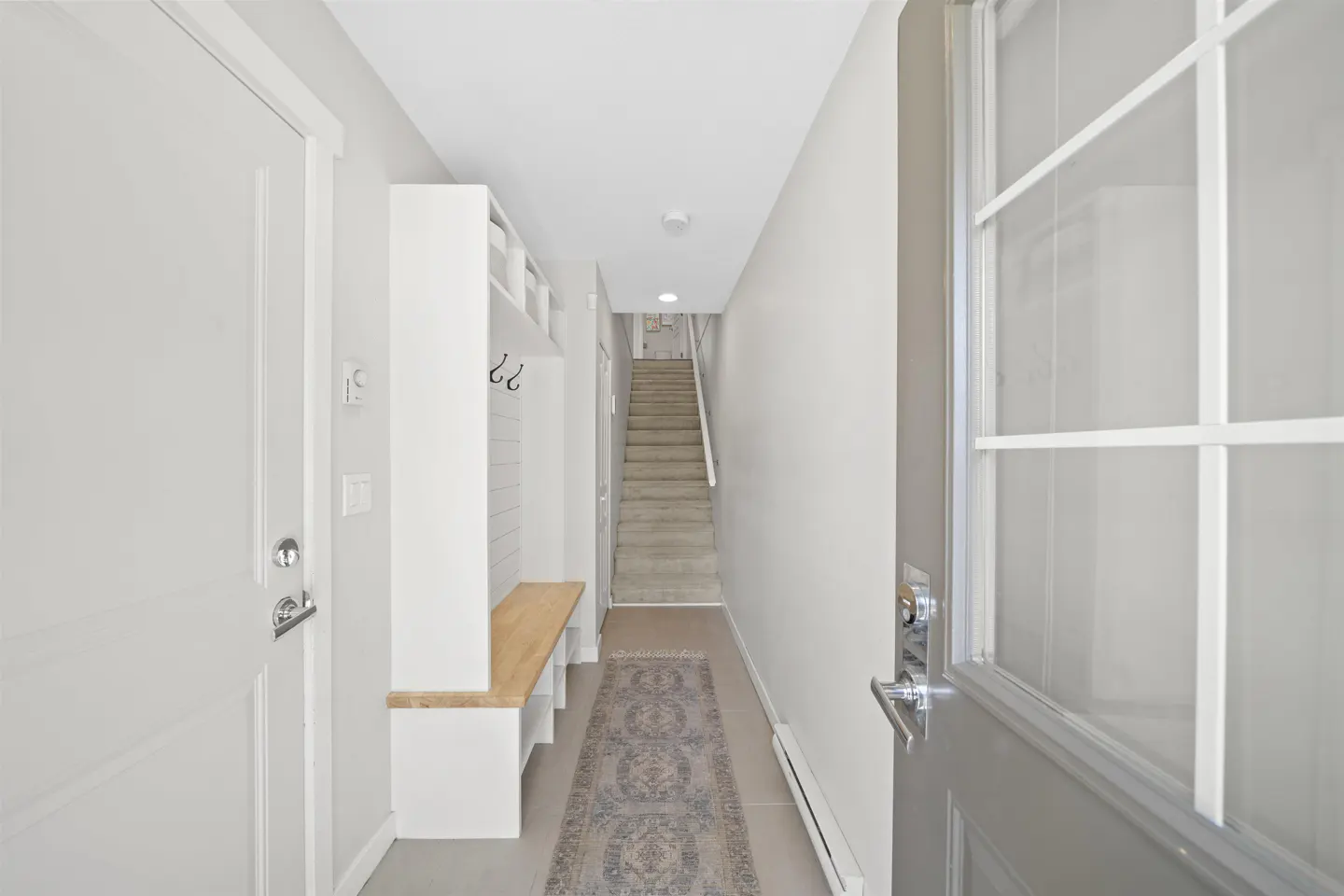 Entryway view with a gray door, white walls, and a built-in bench with hooks. A rug leads to stairs in the background.