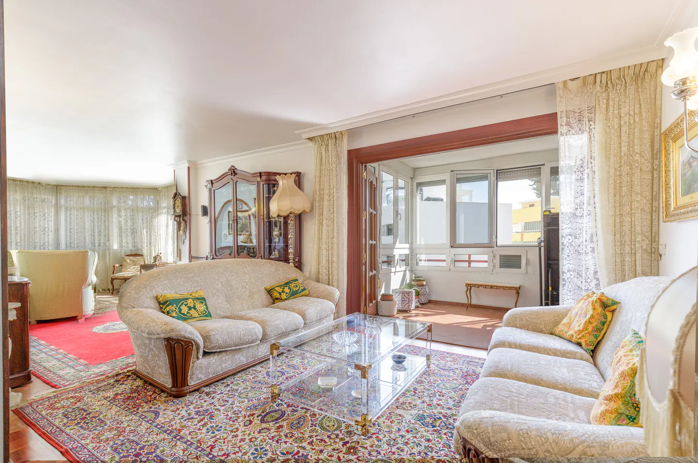 A bright living room with two beige sofas, a glass coffee table, and a colorful patterned rug. A sunroom is visible through an open doorway.