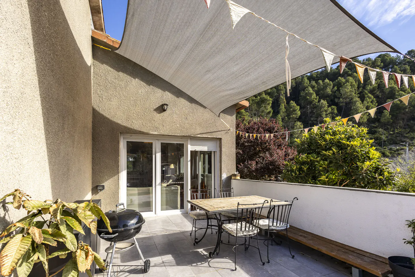 Outdoor patio with a grill, table, and chairs under a sunshade and party flags, with a view of trees.
