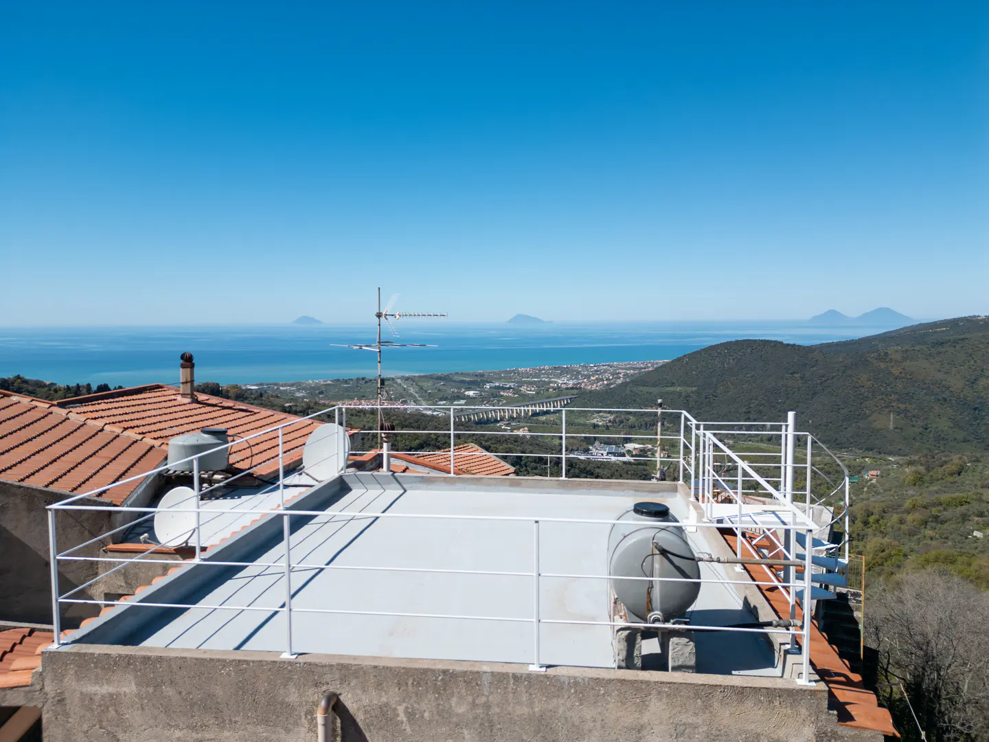 Rooftop view with white railing, gray water tank, and satellite dishes. Distant ocean, islands, and green hills under a clear blue sky.